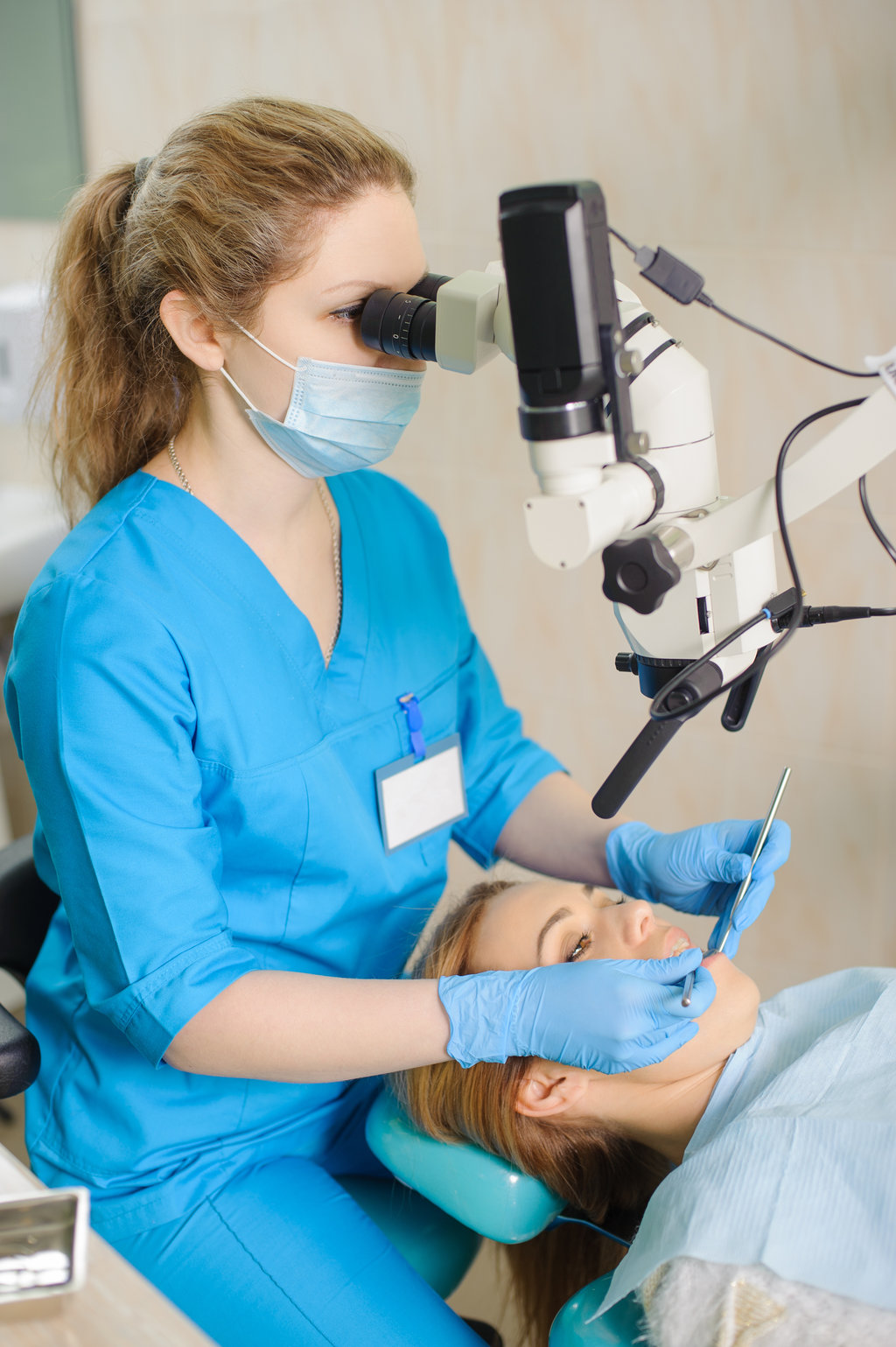 Endodontic Surgery in Allentown, PA 1 Young woman dentist treating root canals using microscope at the dental clinic. Young girl patient lying on dentist chair with open mouth. Dentist wearing mask, gloves and medical suit.