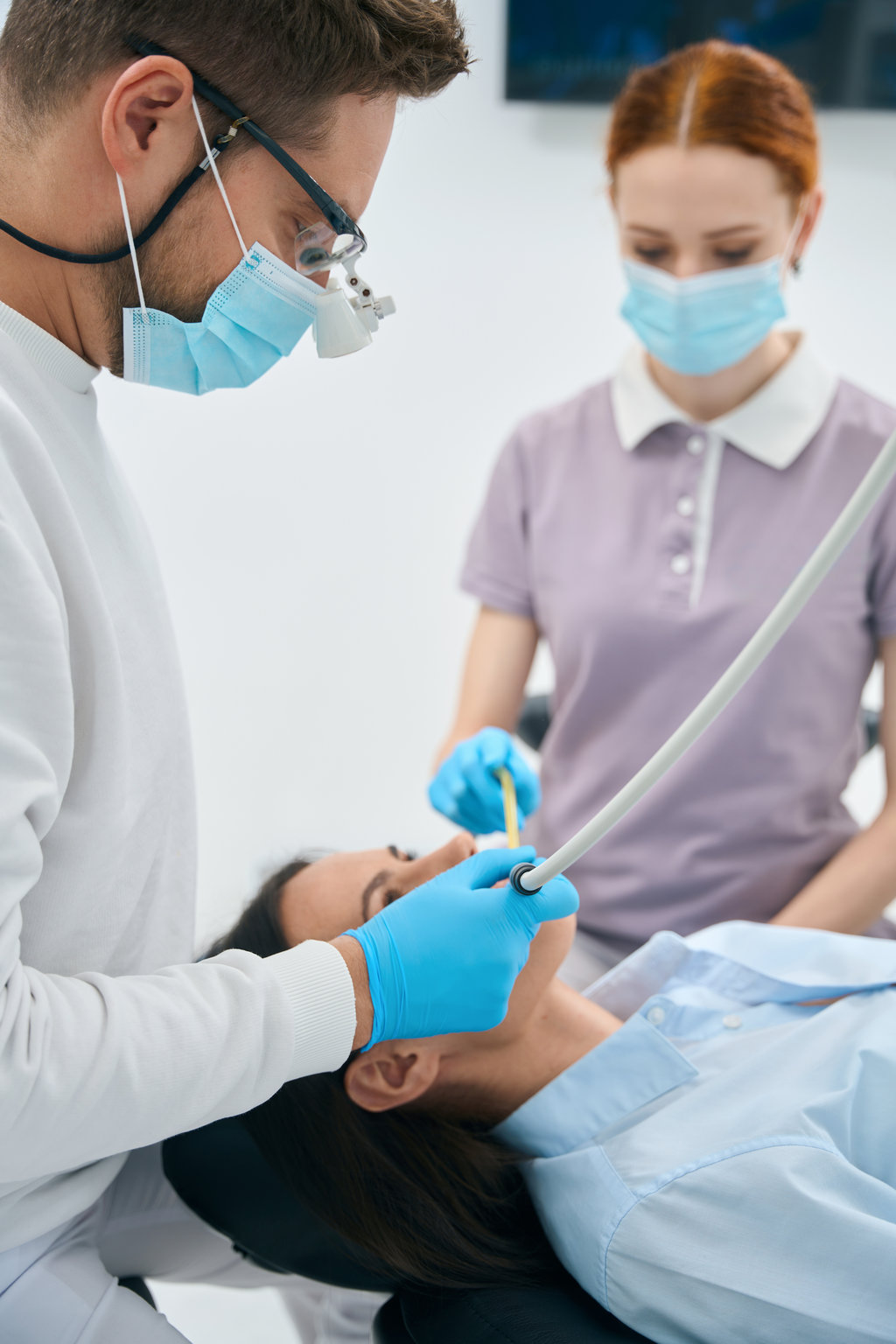 Focused doctor dental technician treating female client teeth, removing tooth decay before filling a cavity, nurse assisting holding suction device