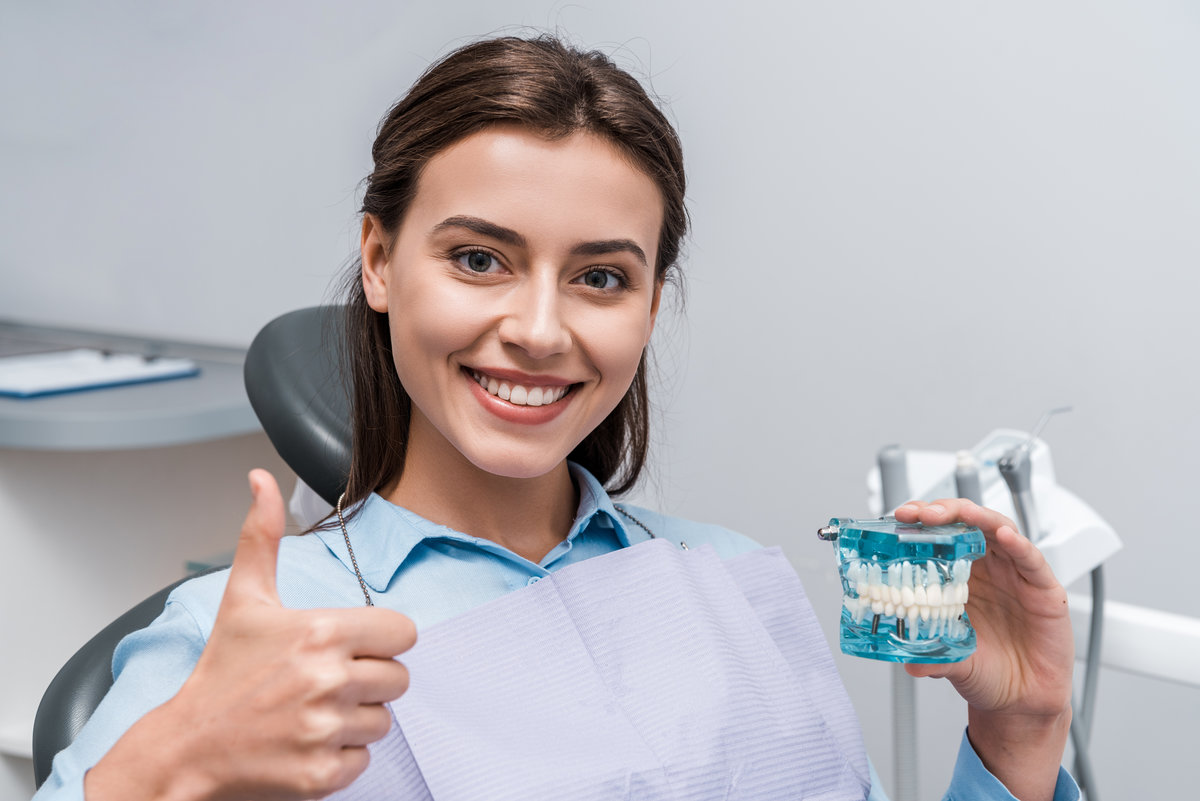 happy woman holding teeth model and showing thumb up in dental clinic